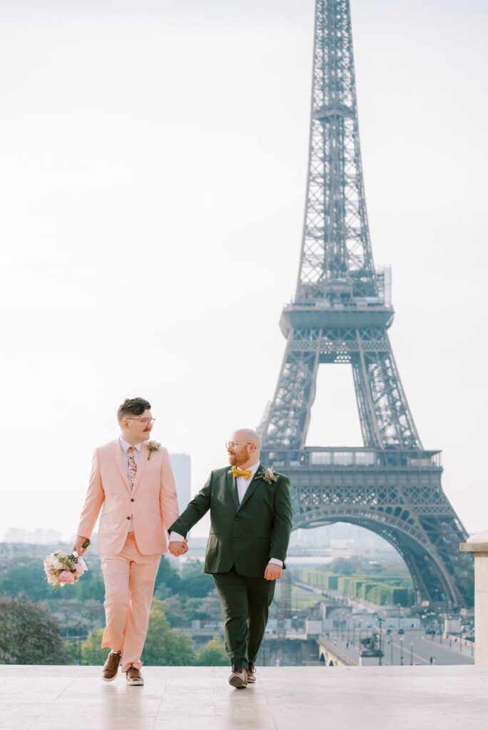 Romantic pastel-toned photo of a gay couple walking together at Trocadero, holding hands and carrying a bouquet with the Eiffel Tower in the background.