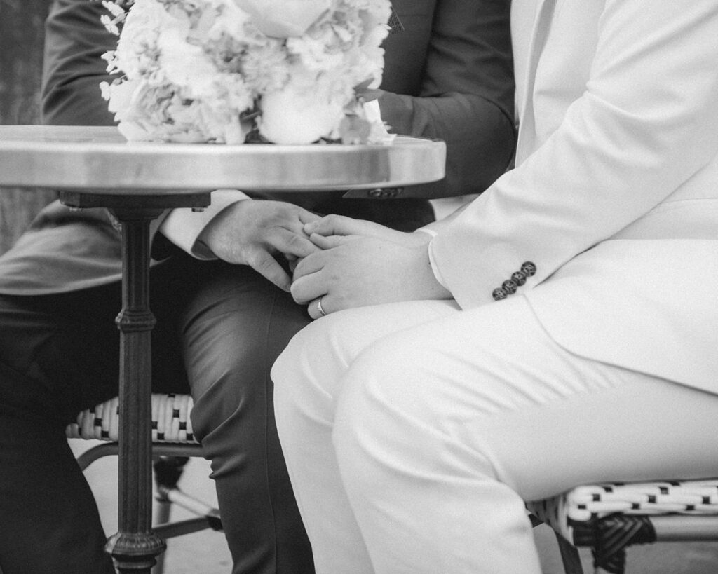 Black and white photography of two men in suits sitting at an outdoor cafe in the 16th arrondissement and holding hands under the table.