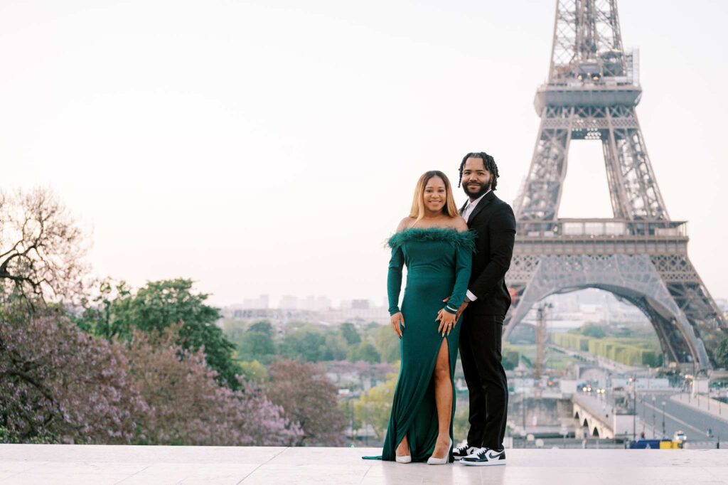 A chic couple dressed formally stands in front of the Eiffel Tower on a clear day, capturing their romantic engagement photoshoot. The woman wears a green gown, and the man is in a black suit with sneakers. Trees and the cityscape add to the enchanting Parisian backdrop.
