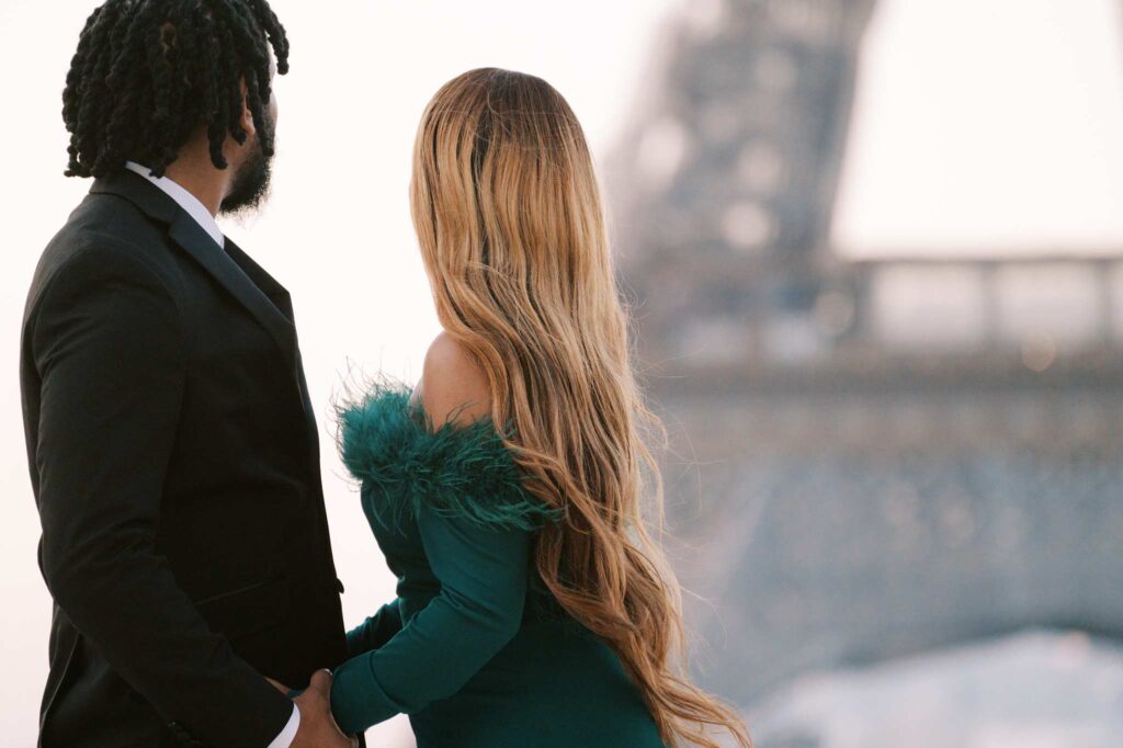 A Black man in a suit and a Black woman in a green dress with long blonde hair stand facing the Eiffel Tower, holding hands during their romantic engagement photoshoot in Paris.