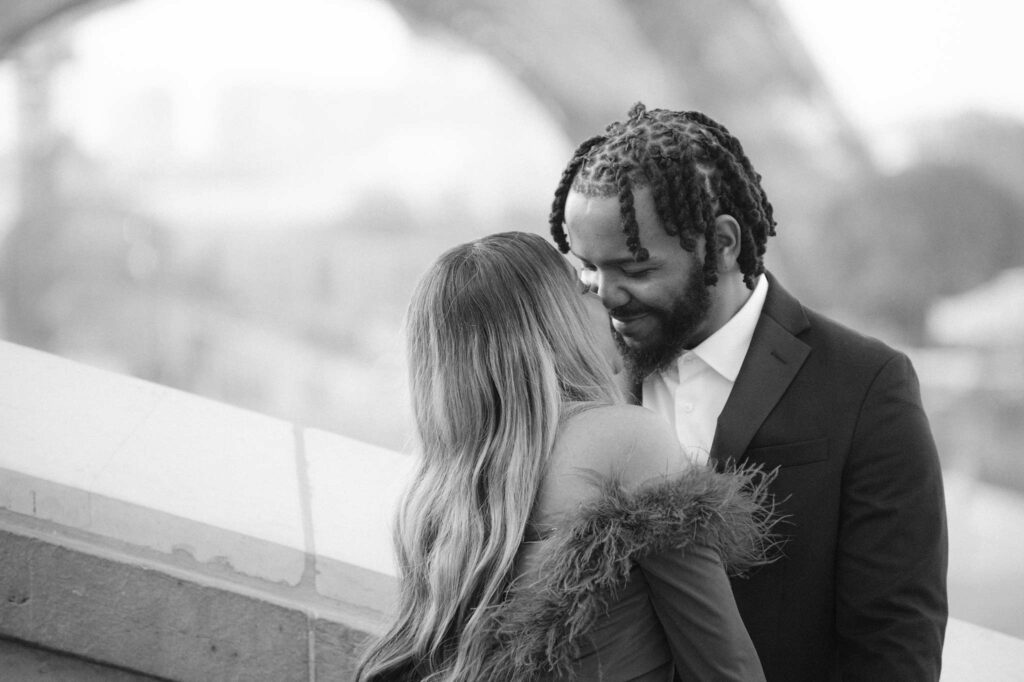 Black and white image of a newly engaged Black couple in Paris during their engagement photo shoot. The man has twists and is wearing a suit and button-down shirt, and the woman has blonde highlights and is wearing an off-the-shoulder dress. They are clearly in love and about to kiss with the Eiffel Tower blurry behind them.