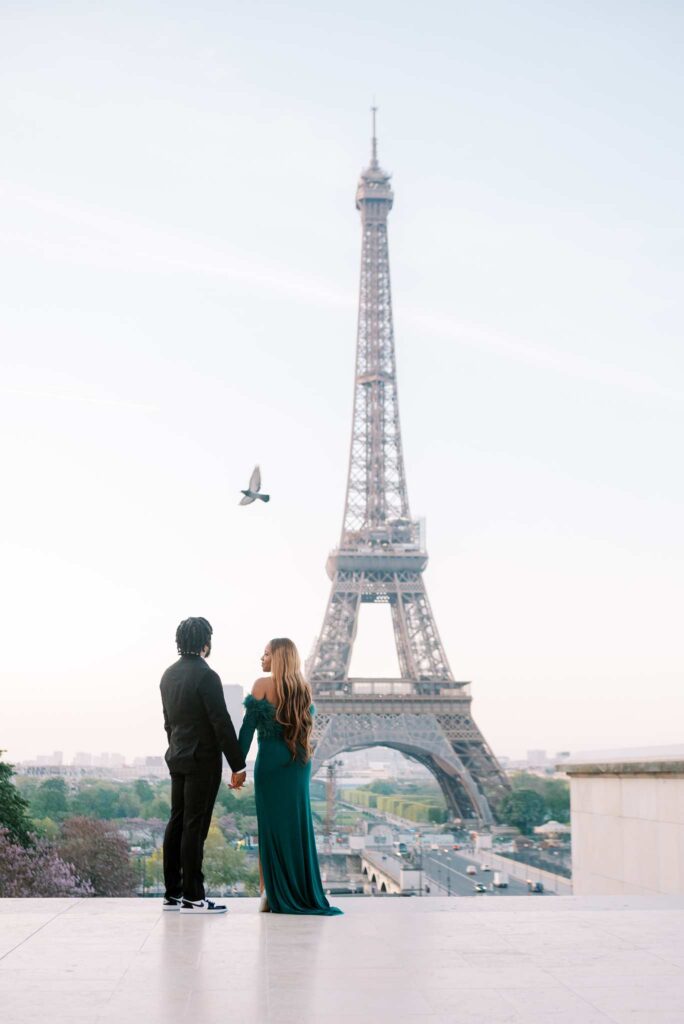 Two people holding hands, facing away from the camera and towards the Eiffel Tower just after sunrise. A bird flies above them. The person on the left wears a dark suit and looks at the tower, as his partner in a long green dress looks towards him.