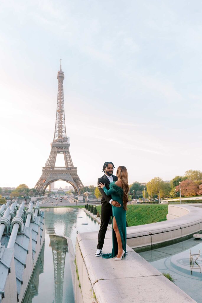 A Black couple in formal attire embraces at the edge of a fountain during their engagement session, with the Eiffel Tower and a park visible in the background.