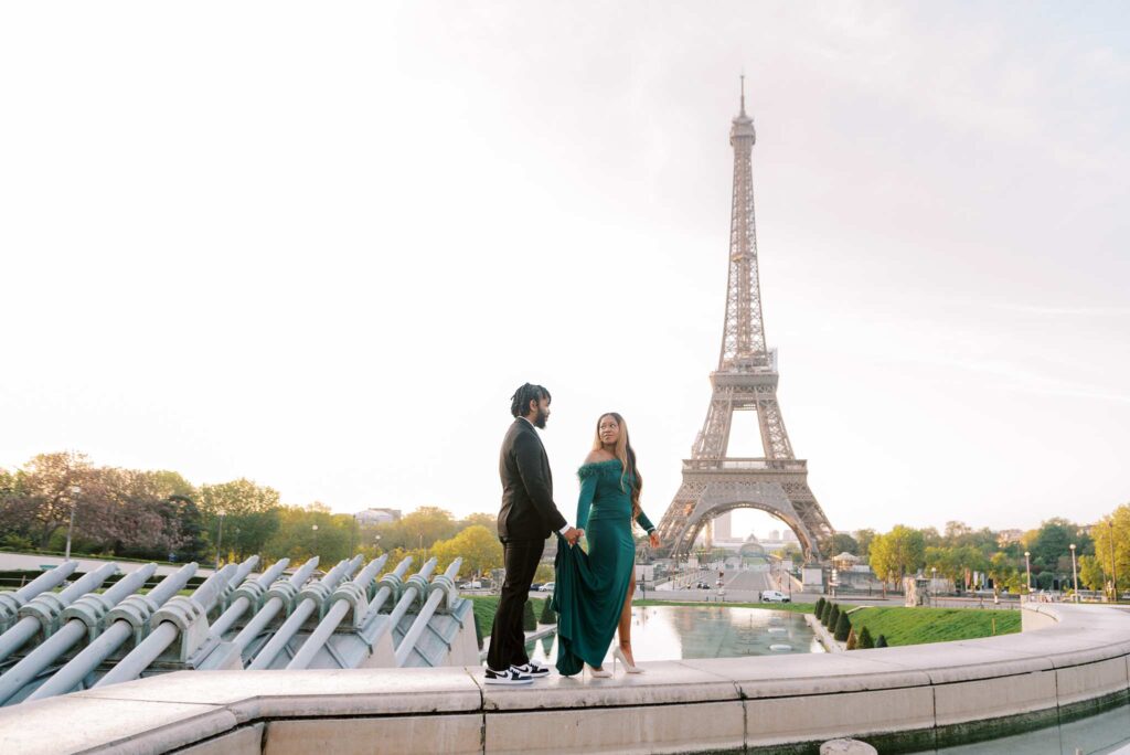 A couple stands on a ledge, looking at each other and holding hands, with the Trocadero fountains and the Eiffel Tower in the background.