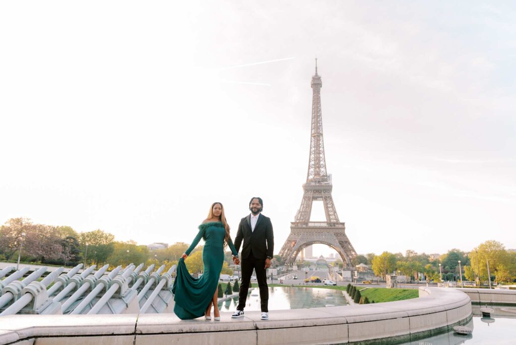 The engaged couple holds hands and looks at the Eiffel Tower with the Trocadero Fountains and the Eiffel Tower behind them.
