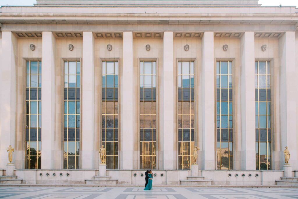 A couple in formal clothing stands in front of a large building at Place du Trocadero with tall columns and gold statues, facing each other in the center of the iconic geometric patterned plaza—a perfect glam setting for their romantic engagement photoshoot in Paris.