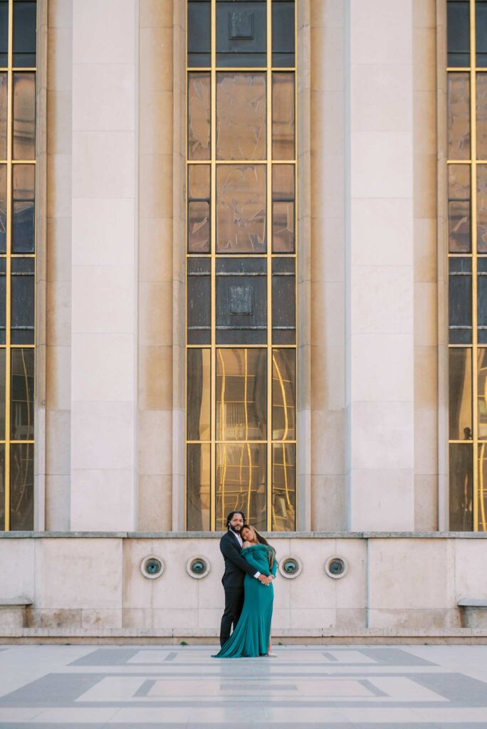 A chic couple holds each other in their arms, standing in front of a large Parisian building with tall windows and gold accents at Place du Trocadero. The woman is in an emerald gown and the man is dressed in a black suit for their engagement photoshoot.