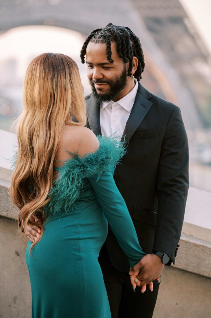 An elegant couple holds hands and faces each other romantically with the blurred iconic monuments of the Eiffel Tower in the background.