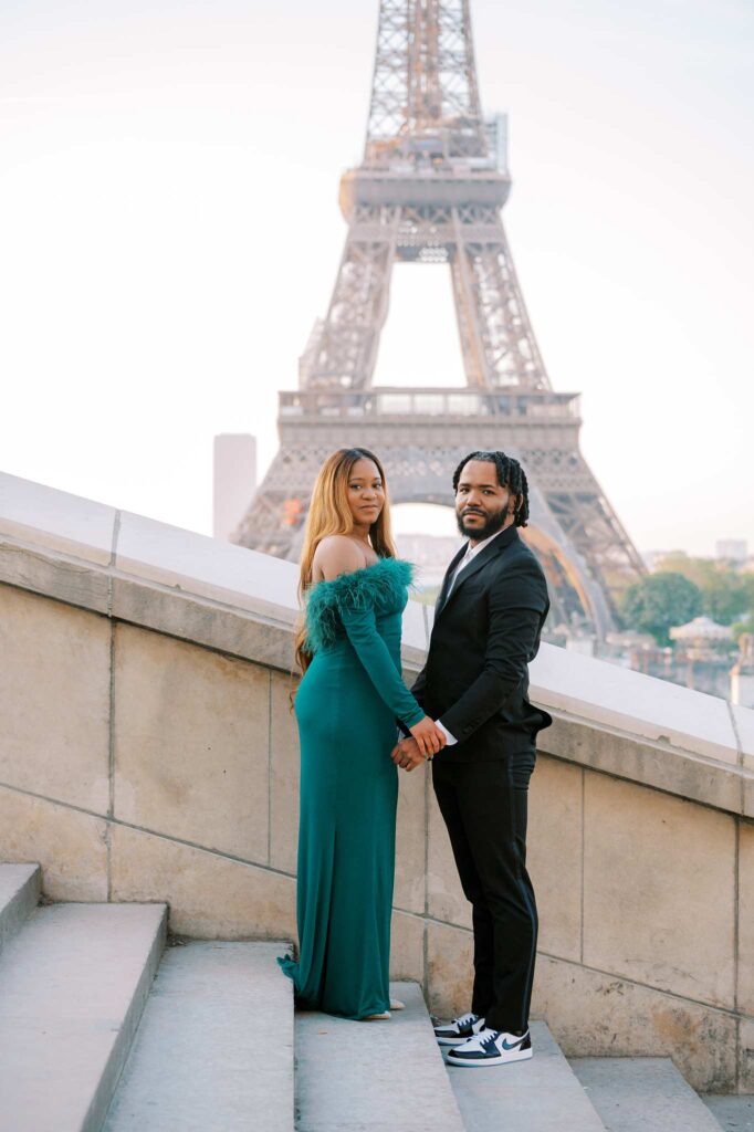 The couple stands on stairs, holding hands and looking at the camera with the Eiffel Tower in the background.