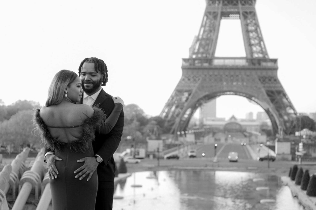 A gorgeous Black couple is holding each other while standing in front of the Eiffel Tower and the Trocadero fountain during their romantic engagement photoshoot. They're in a slow dancing position: the man faces the camera and looks at his fiancé with his hands on the small of her back. His fiancée has her arms on his shoulders and faces away from the camera and looks to the side. Her long straight hair is swept off to the side as well, showing off the back of her dress as well as her earrings. The scene is timelessly captured in black and white.