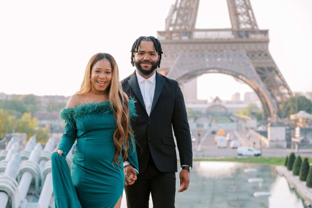 The newly engaged couple holds hands and smiling at the camera, with a fountain and the Eiffel Tower behind them.