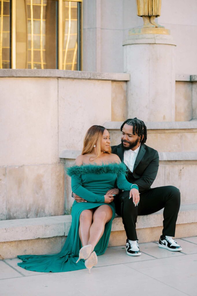A couple dressed up in formal attire sits on a stone ledge at Place du Trocadero. The woman wears an off-the-shoulder green dress, and the man wears a black suit with sneakers. Smiling and looking at each other, they exude a romantic Parisian charm perfect for an engagement photoshoot.
