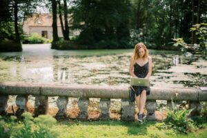 A woman in a dress sits on a stone railing by a pond, working on a laptop in a lush, green outdoor setting with a house and trees in the background, probably reading up on some Lightroom tips to enhance her photography skills.