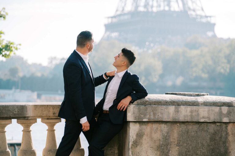 Two men stand in front of a stone railing on a bridge with the Eiffel Tower in the background. One man has his hand on the other's shoulder, and he looks toward the tower as his partner looks up at him.