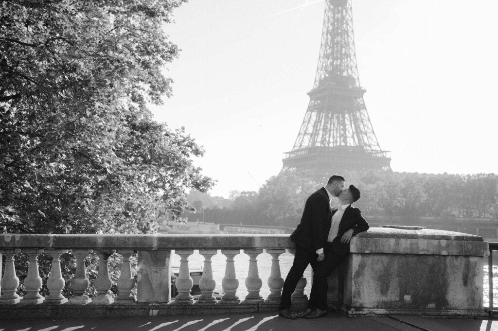 A gay couple shares a kiss while leaning against a railing, with the Eiffel Tower visible in the background, capturing a romantic moment during their photo shoot in Paris.