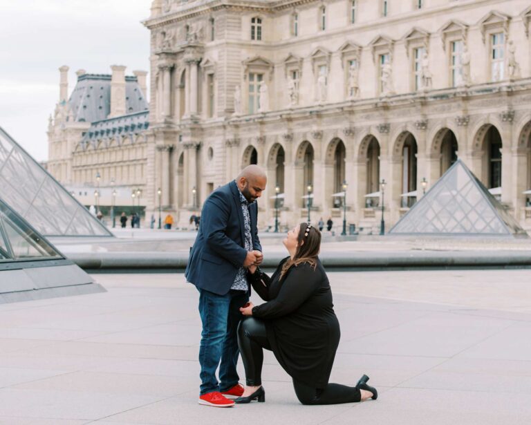 A woman kneels while holding a ring, proposing to a man who stands smiling and looking down at her. This surprise proposal unfolds in front of the Louvre Museum with its iconic glass pyramids and iconic Parisian facades in the background. Both are dressed casually, the man in a blazer and jeans, and the woman in black leggings and a black dress.