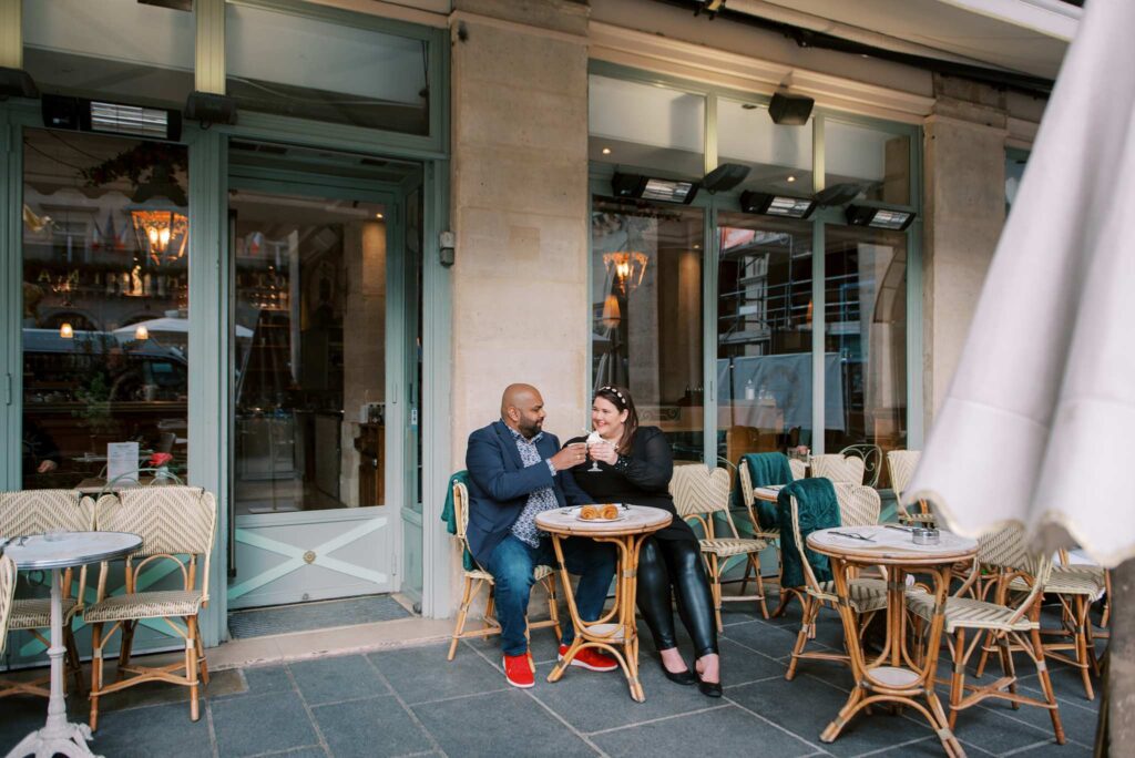 A man and a woman sit at a small round table outside a cozy Paris café, enjoying croissants and hot drinks. They clink glasses in a toast following the woman proposing to her soon-to-be-husband.