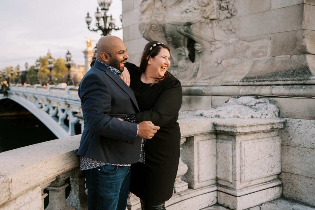 An Indian man wearing a navy blazer and a white woman in a black dress stand together on the ornate Pont Alexandre III. Ornate street lamps line the bridge in the background as they lean against the stone railing, sharing a moment of laughter and looking off into the distance.