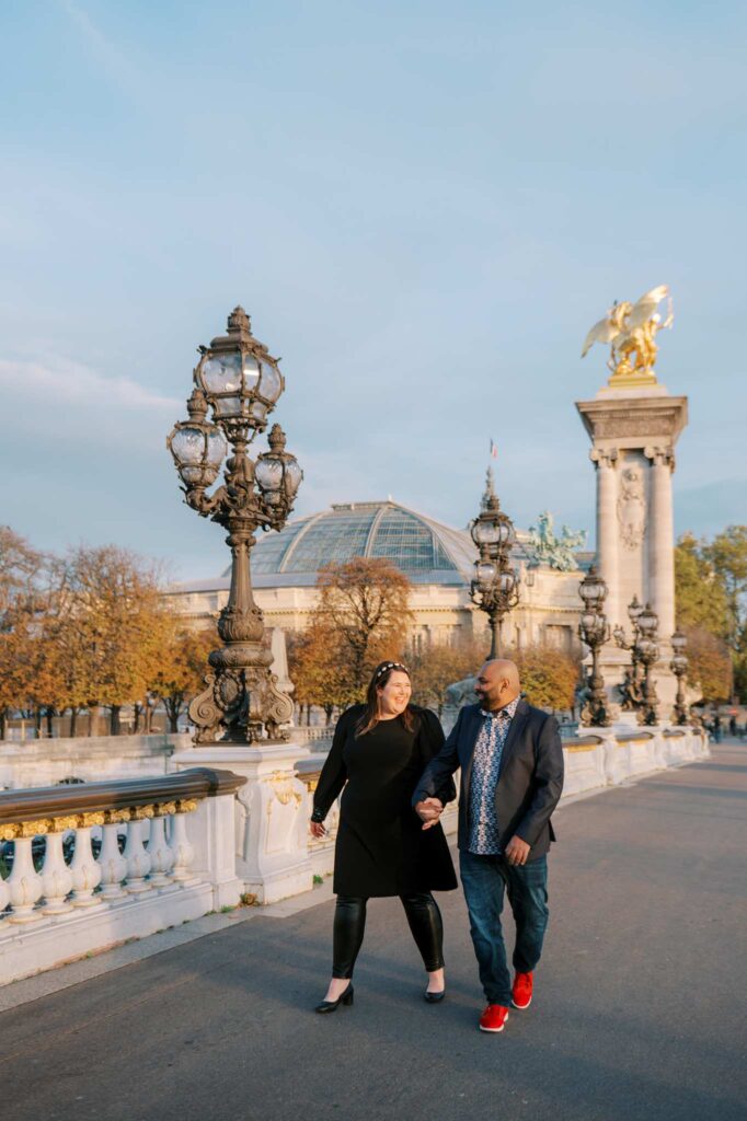 A couple holding hands and walking on the beautiful Pont Alexandre III with large light posts and a golden statue in the background. Trees and the glass dome of the Grand Palais are visible behind them.
