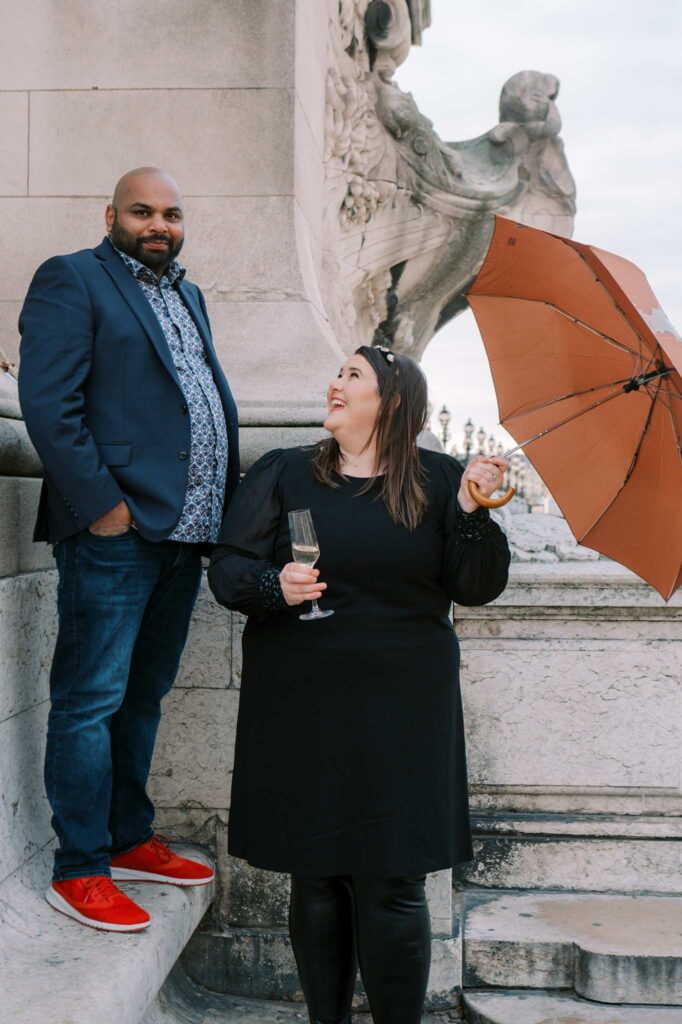 Two people stand on stone steps at the edge of a bridge. On the left, a man in a blue blazer, blue jeans, and red shoes looks at the camera. On the right, a woman in a black dress holds an brown umbrella and a half-full champagne glass, smiling up at the man.