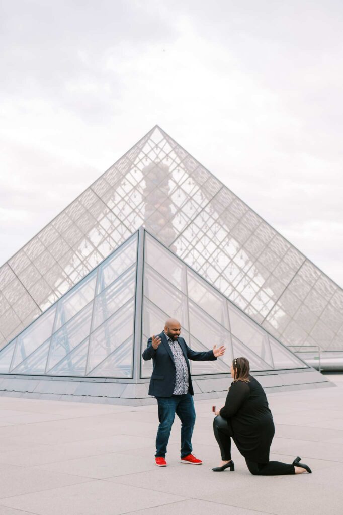 A woman kneels for a surprise proposal in front of the Louvre Pyramid in Paris. Her partner has just turned around, and he looks surprised and excited with arms open wide. The glass and metal structure of two pyramids forms a striking backdrop, adding to the magic of the moment.
