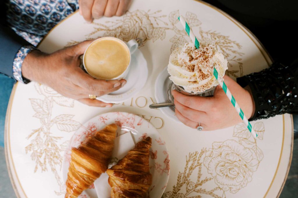 A top-down view shows two hands resting on a Parisian cafe table. One person holds a cup of coffee, while the other holds a hot chocolate topped with whipped cream, with two croissants on a floral plate waiting to be eaten. There is an engagement ring on the hand of each partner.