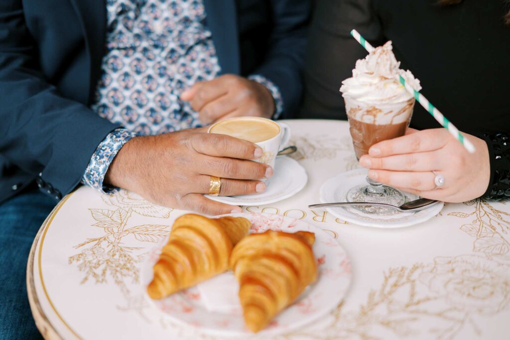 Close-up of two people sitting at a table in a cozy café, each holding a drink. One person has a cup of coffee, and the other has a whipped cream-topped hot chocolate drink. On the table are two croissants. They are both wearing engagement rings after the surprise proposal that she carefully planned.