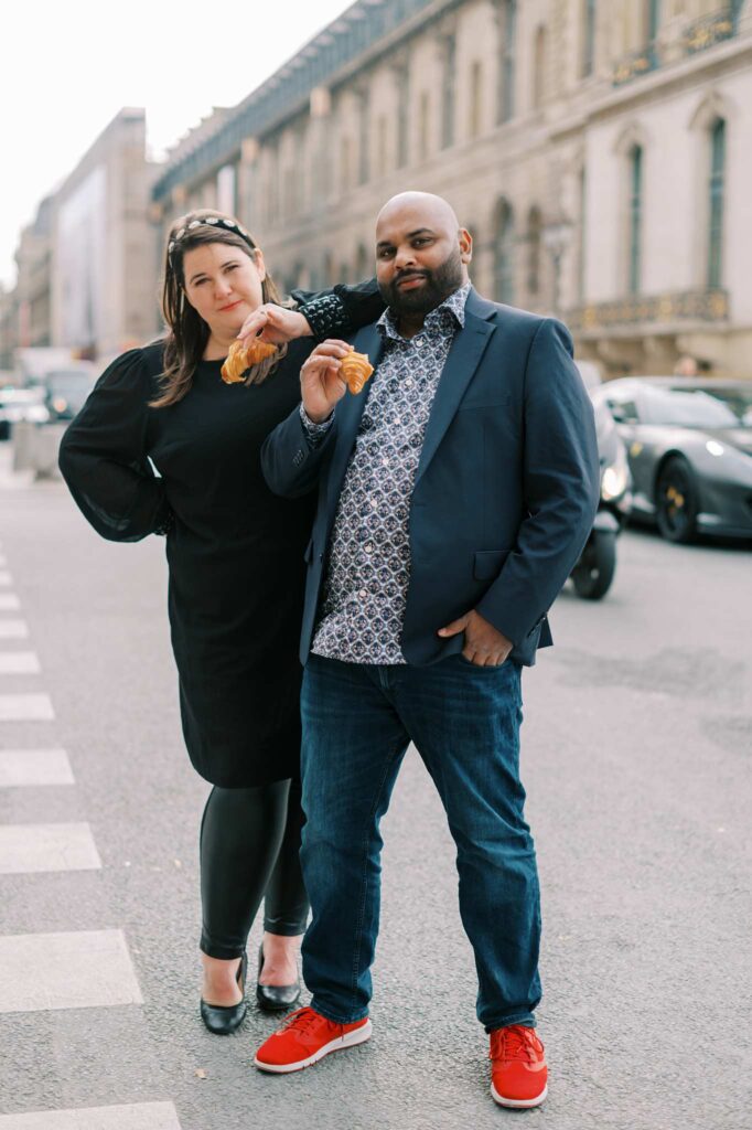 A man and a woman pose on a Paris street holding croissants at the end of their photoshoot. Their vibe is a little bit serious, a little bit "too cool for school."
