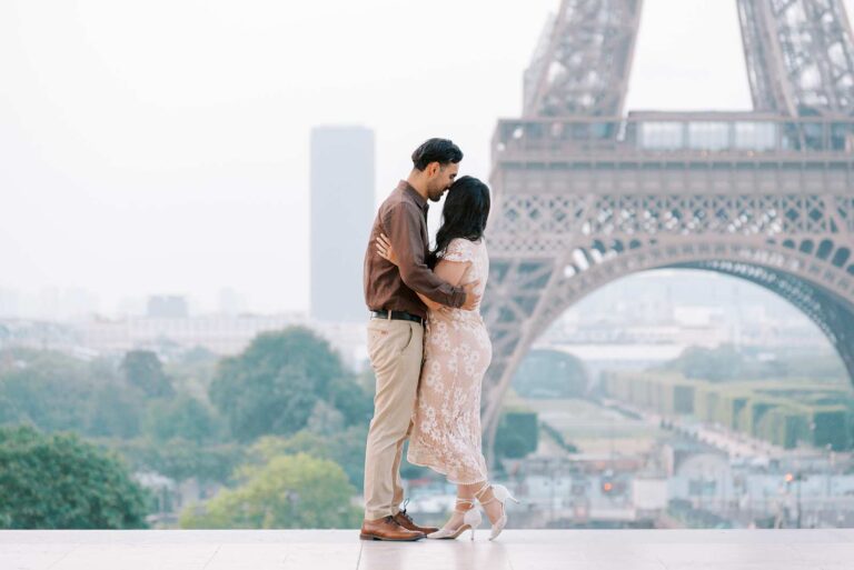 A couple stands close together and holds each other at Place du Trocadero with the Eiffel Tower in the background. The woman wears a light-colored lace dress, and the man is dressed in a brown button-up shirt and beige pants.