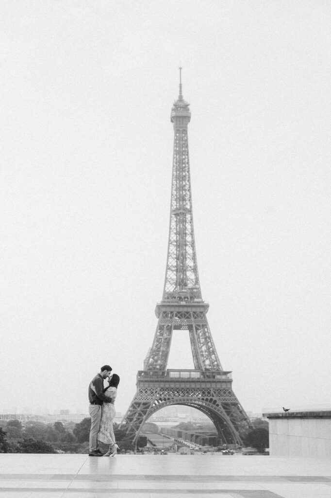 A couple kisses at Place du Trocadero, with the full Eiffel Tower behind them, during their 4th anniversary photo session in Paris, France. The black and white photo captures the romantic moment with the iconic monument prominently standing in the background. It's a serene and slightly foggy early morning.