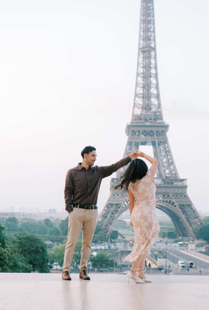 A man and a woman dance in front of the Eiffel Tower in Paris during their anniversary photo session. The man, wearing a dark long-sleeve shirt and beige pants, twirls the woman in her white lacy dress and high heels.
