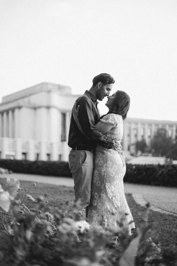 Black and white image of a couple embracing tenderly outdoors. They are in the Trocadero gardens, with a flower bed in the foreground and Place du Trocadero in the distance behind them.