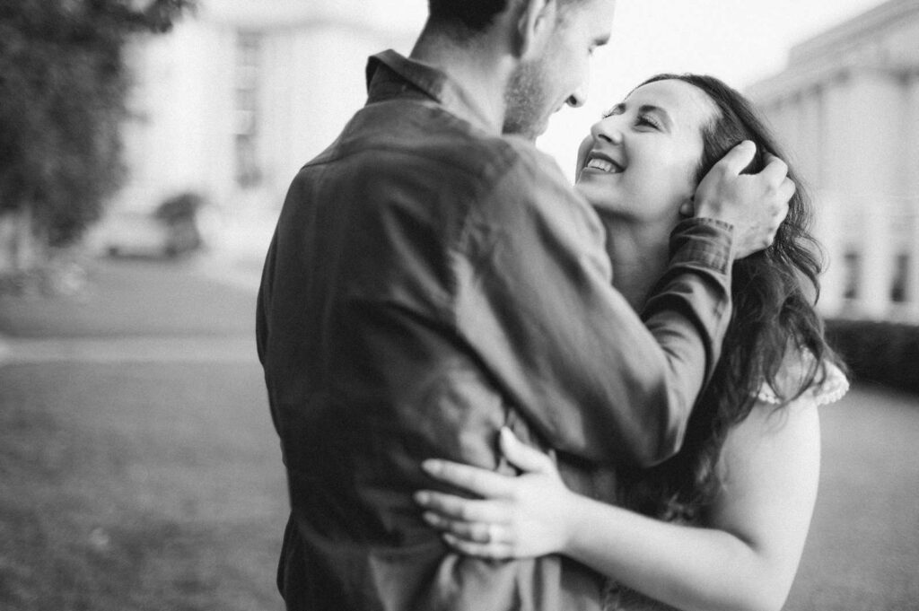 A black and white photo of a couple outdoors in Paris, smiling warmly at each other. The man gently holds the woman’s face as he sweeps her hair out of her eyes. In the background are the blurred buildings and gardens of Trocadero.