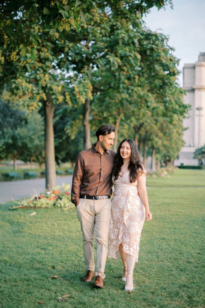 A couple walks with their arms around each other through the grass near Place du Trocadero, with trees and a pathway behind them.