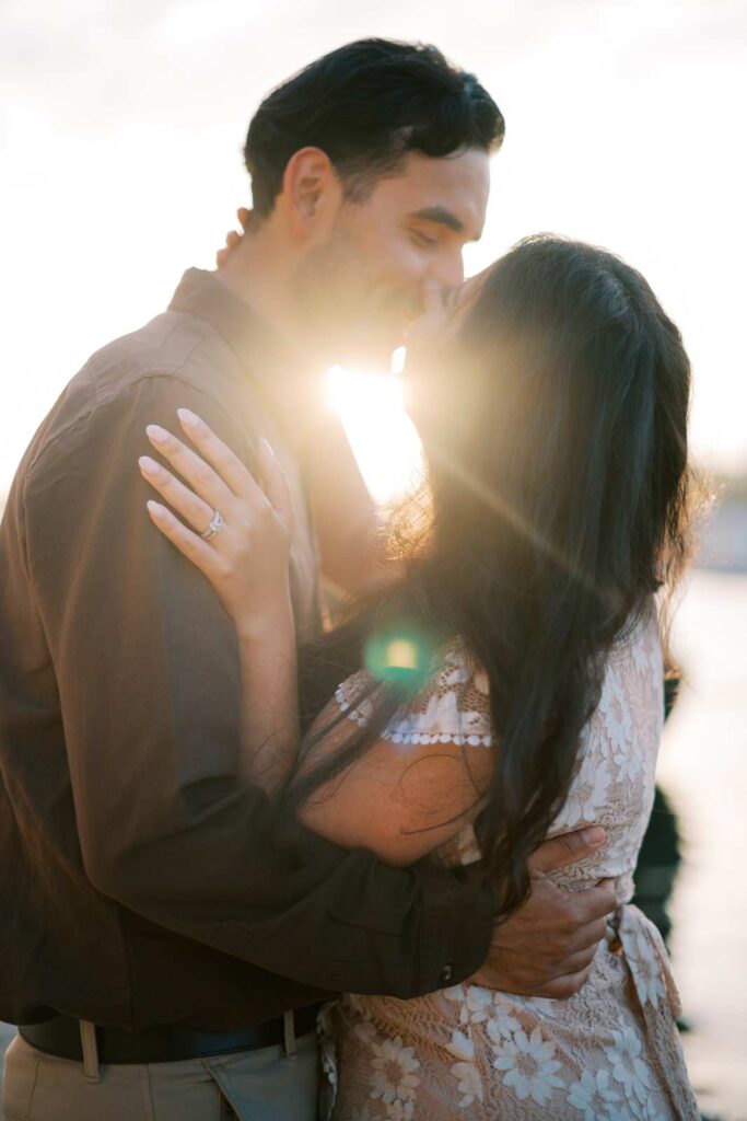 Glowy, backlit, romantic photo of a couple sharing an intimate kiss as the sun rises over the Seine River in Paris. The man's arms are wrapped around the woman's waist, and she has one hand on his chest, showcasing her engagement ring and wedding ring. The warm sunlight creates a ethereal glow around them, perfect for their 4th anniversary celebration photo session.