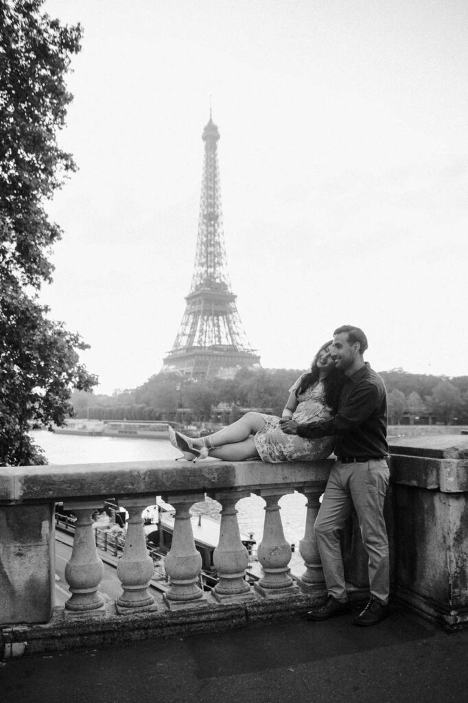 A black-and-white photo shows a woman sitting on the stone railing of the Bir-Hakeim bridge and leaning on her husband, while he stands beside her with his arms around her. The Seine river and Eiffel Tower are in the background.