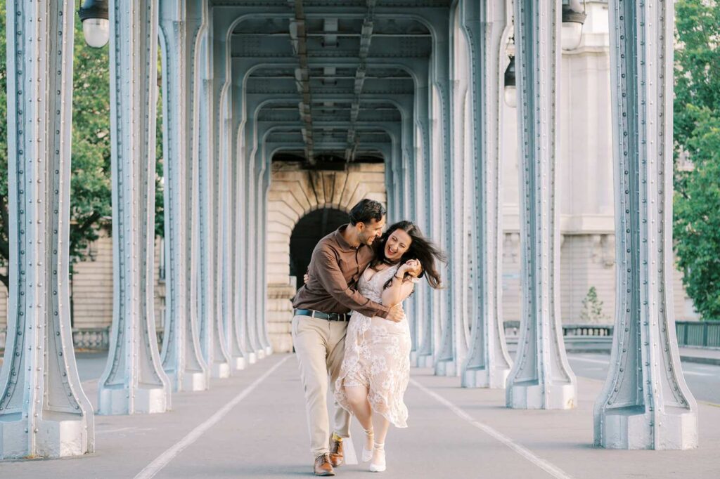 A couple walks while holding each other close, down a path surrounded by an archway of metal beams on the Bir-Hakeim bridge. It's a candid moment and they're both laughing, and the woman's hair is blowing in the wind.