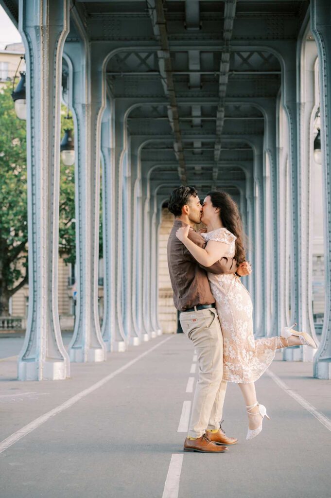 A man lifts his wife off the ground on the Bir-Hakeim bridge as they share a romantic kiss in Paris during their anniversary photo shoot. They're surrounded by the modern and architectural metal columns of the bridge and they're totally alone.