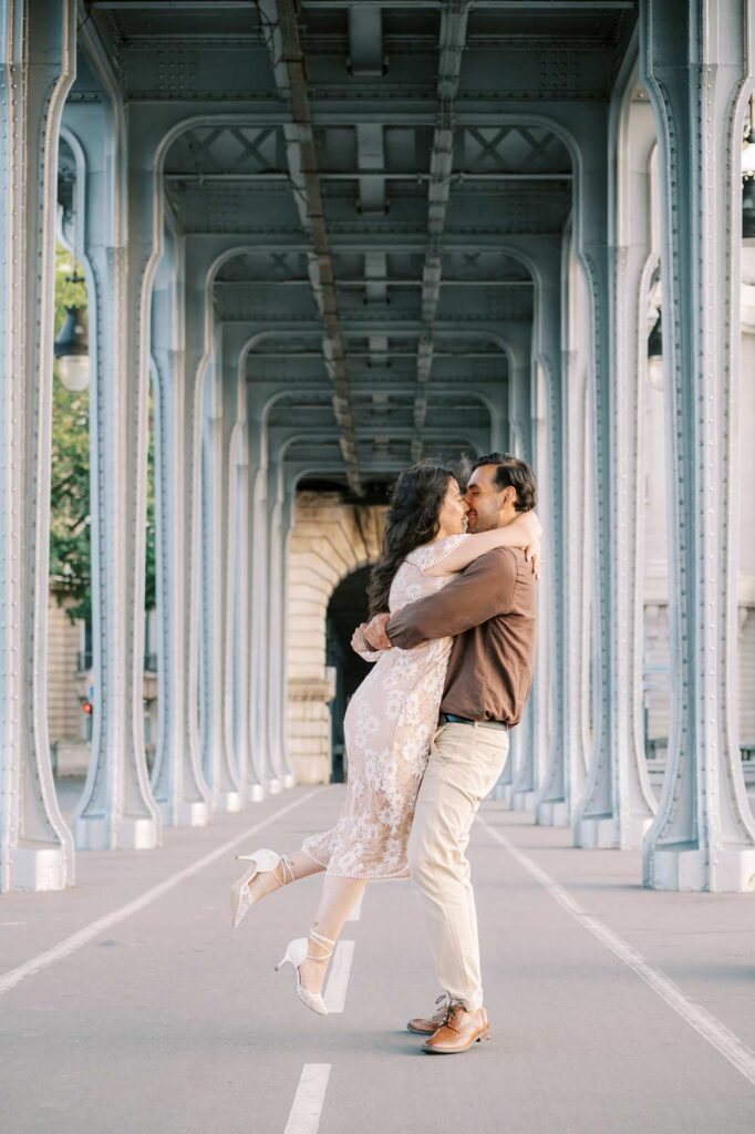 A man lifts his wife off the ground as they kiss on the Bir-Hakeim bridge, surrounded by rows of the bridge's modern metal columns.