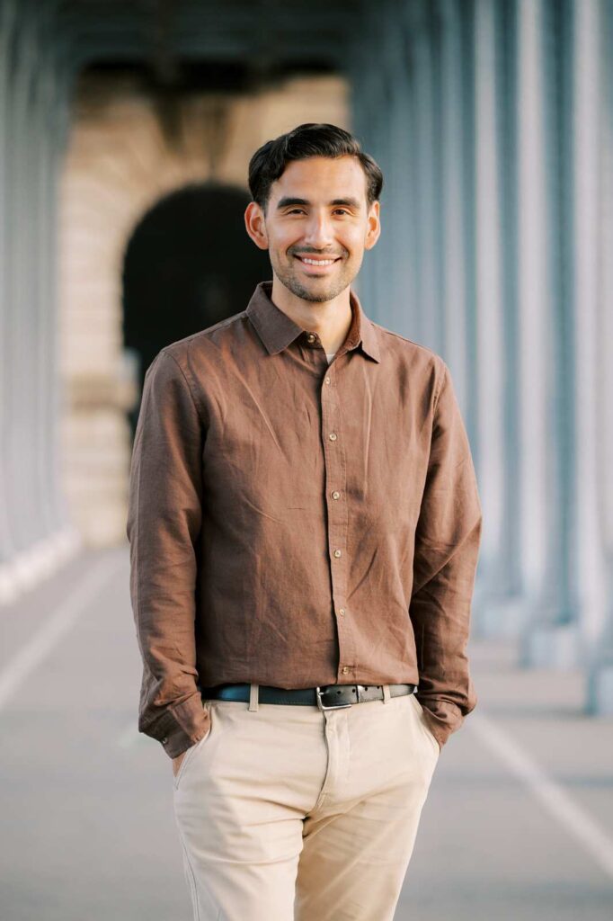 Portrait of a man with hands in his pockets smiling at the camera during a photo session. The background features a metal architectural structure with arches, part of the Bir-Hakeim bridge in Paris.