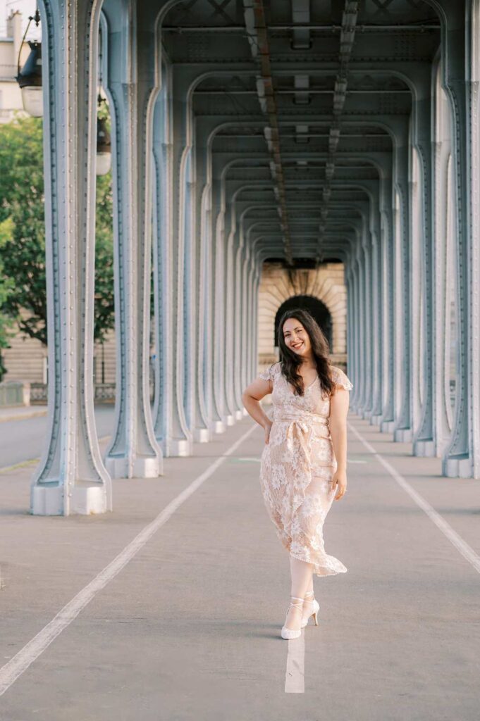 A woman in a cream floral dress smiles for the camera on a pathway beneath a series of metallic arches. She has long, dark hair and is wearing white shoes.