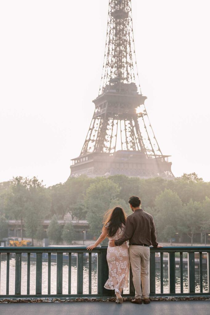 A couple stands on the Bir Hakeim bridge, facing the Seine River and the Eiffel Tower in the background during a serene early-morning anniversary photo shoot in Paris.