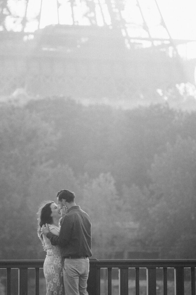 Hazy black and white photo of a couple dancing on a bridge and gazing into each other's eyes. The haze creates a romantic ambiance, and the Eiffel Tower and trees in the background take up most of the framing of this photo.