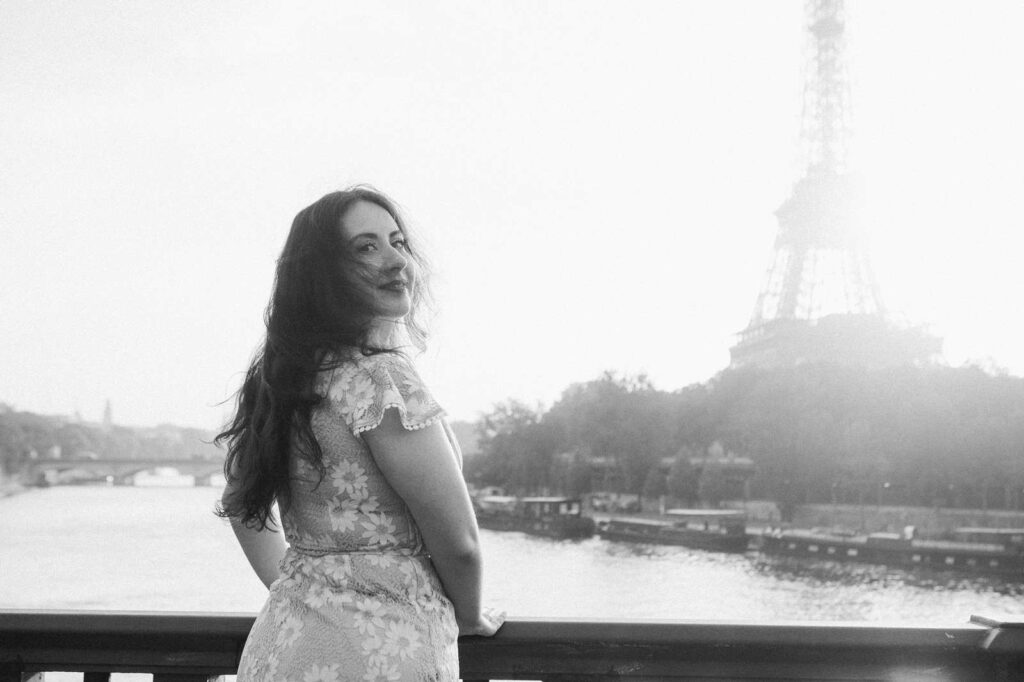 A woman with long hair stands by a railing overlooking the Seine river, and looks back over her shoulder with a smile. The Eiffel Tower is visible in the background.