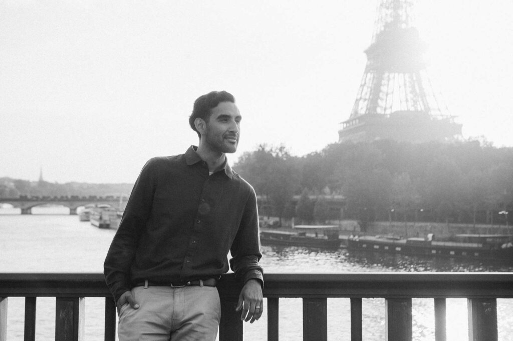 A man leans against a railing on a bridge in Paris, with the Seine and Eiffel Tower in the background.