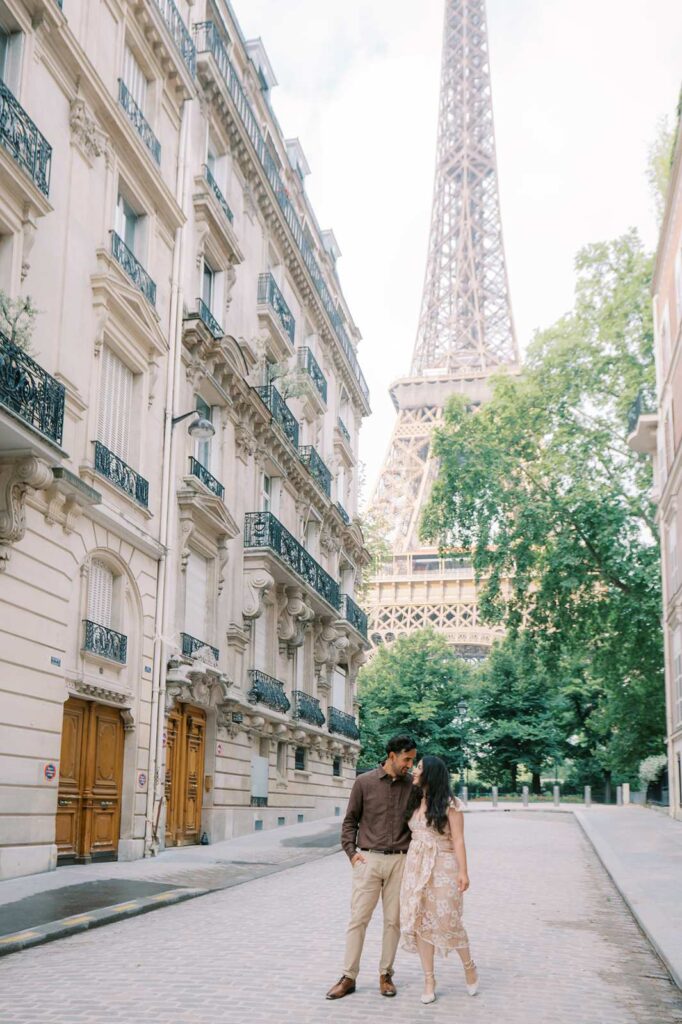 A couple walks hand in hand on the cobblestones at rue de l'Université during their anniversary photo shoot. The Eiffel Tower stands tall in the distance amid lush green trees, and historic Parisian architecture lines the side of the street. The sky is slightly cloudy, creating a soft, romantic atmosphere.