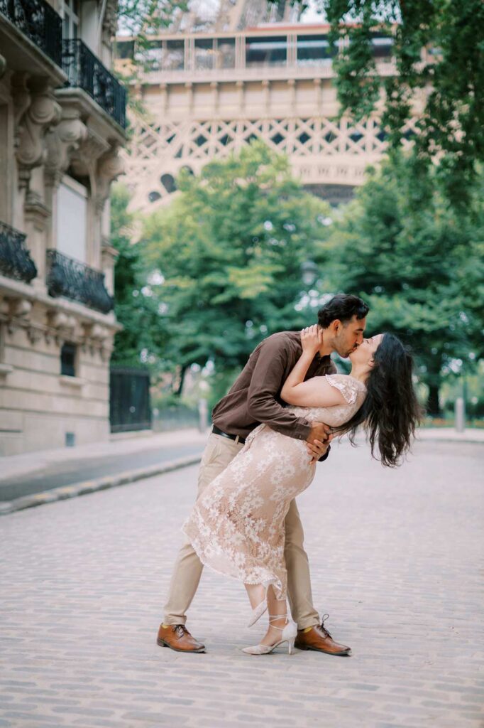 A couple shares a romantic kiss on a cobblestone street with the Eiffel Tower in the background during their 4th anniversary photo session. The man is dipping the woman slightly, and on either side of them are lush green trees and the elegant buildings of rue de l'Université.