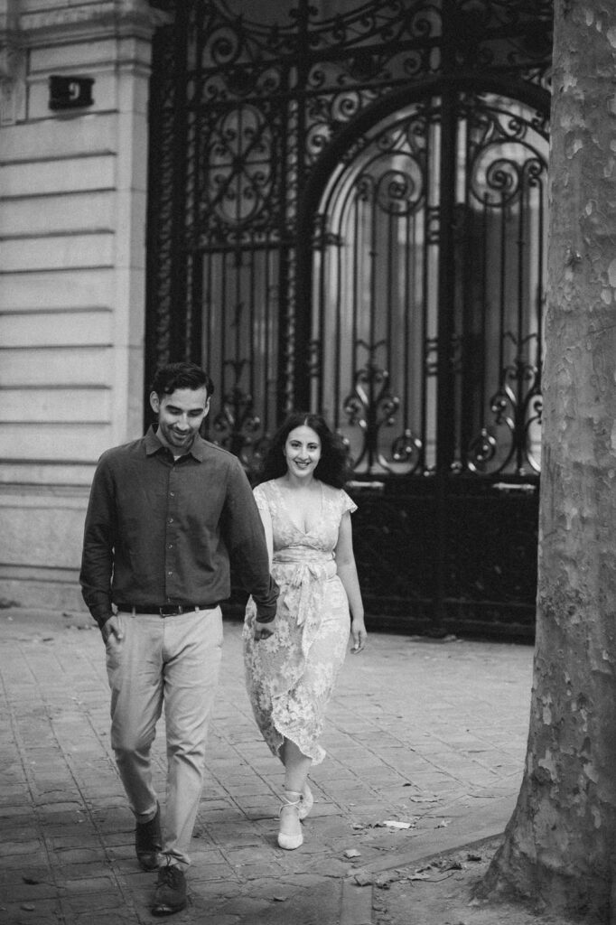 A dark black-and-white photo of a smiling couple walking hand-in-hand away from an ornate wrought-iron gate in Paris.