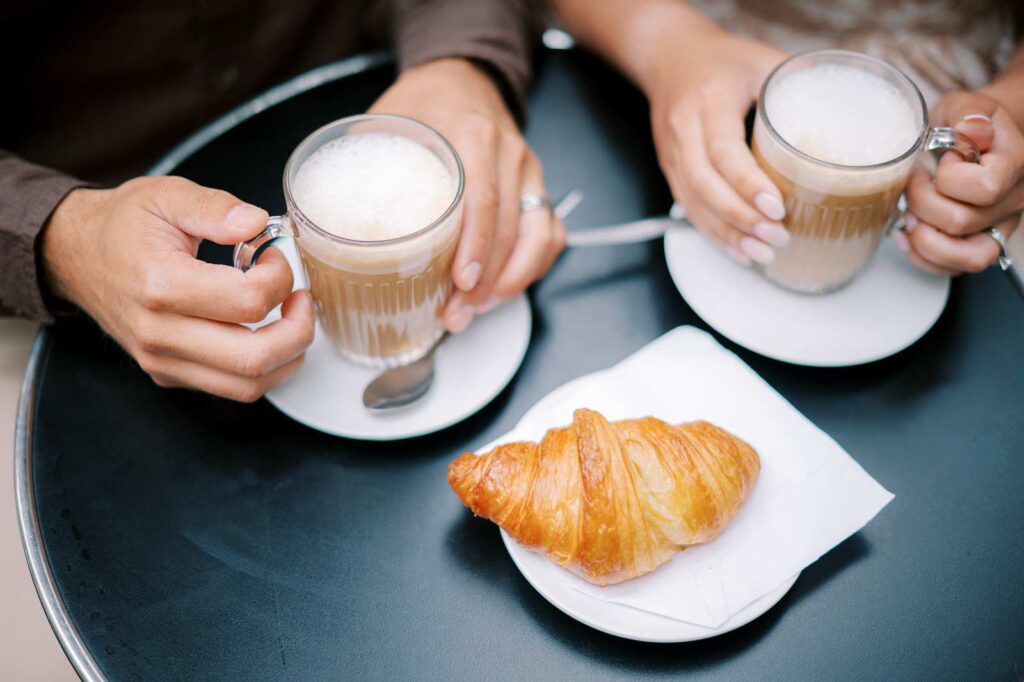 Top-down view of two people's hands holding lattes on a round black table. A croissant on a white napkin sits on a small plate between them, making for a cozy coffee date at the end of their 4th anniversary photo session.
