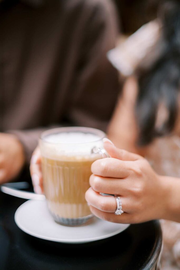 A person with a diamond ring on their finger holds a clear glass mug filled with a latte.